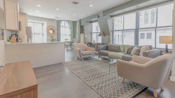 A modern living room with a sofa, armchairs, and a coffee table. There is a kitchen area with cabinets and a countertop. Large windows provide natural light and views of the city. - Luxurious 1BR in FIDI Boston | Close to Everything (Boston)
