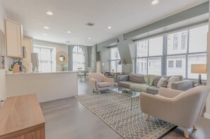 Een moderne woonkamer met een bank, fauteuils en een salontafel. Er is een keuken met kasten en een aanrecht. Grote ramen zorgen voor natuurlijk licht en uitzicht op de stad.