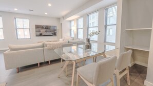 Un salon moderne avec une table à manger en verre, des canapés beige et une grande fenêtre équipée de stores.