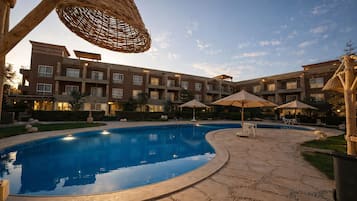 A swimming pool with lounge chairs and umbrellas, surrounded by a courtyard with a multi-story building.