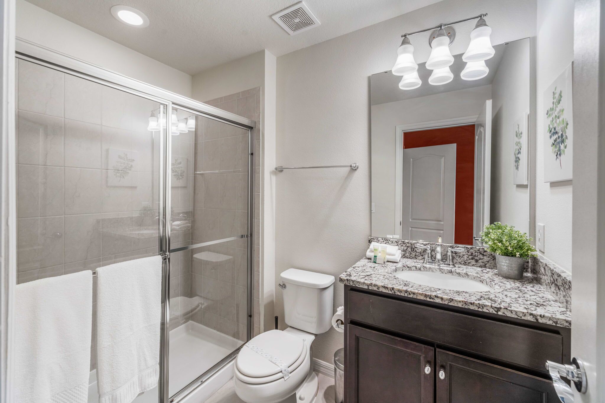 Un baño moderno con ducha de vidrio, inodoro blanco, encimera de granito y espejo con luces arriba.