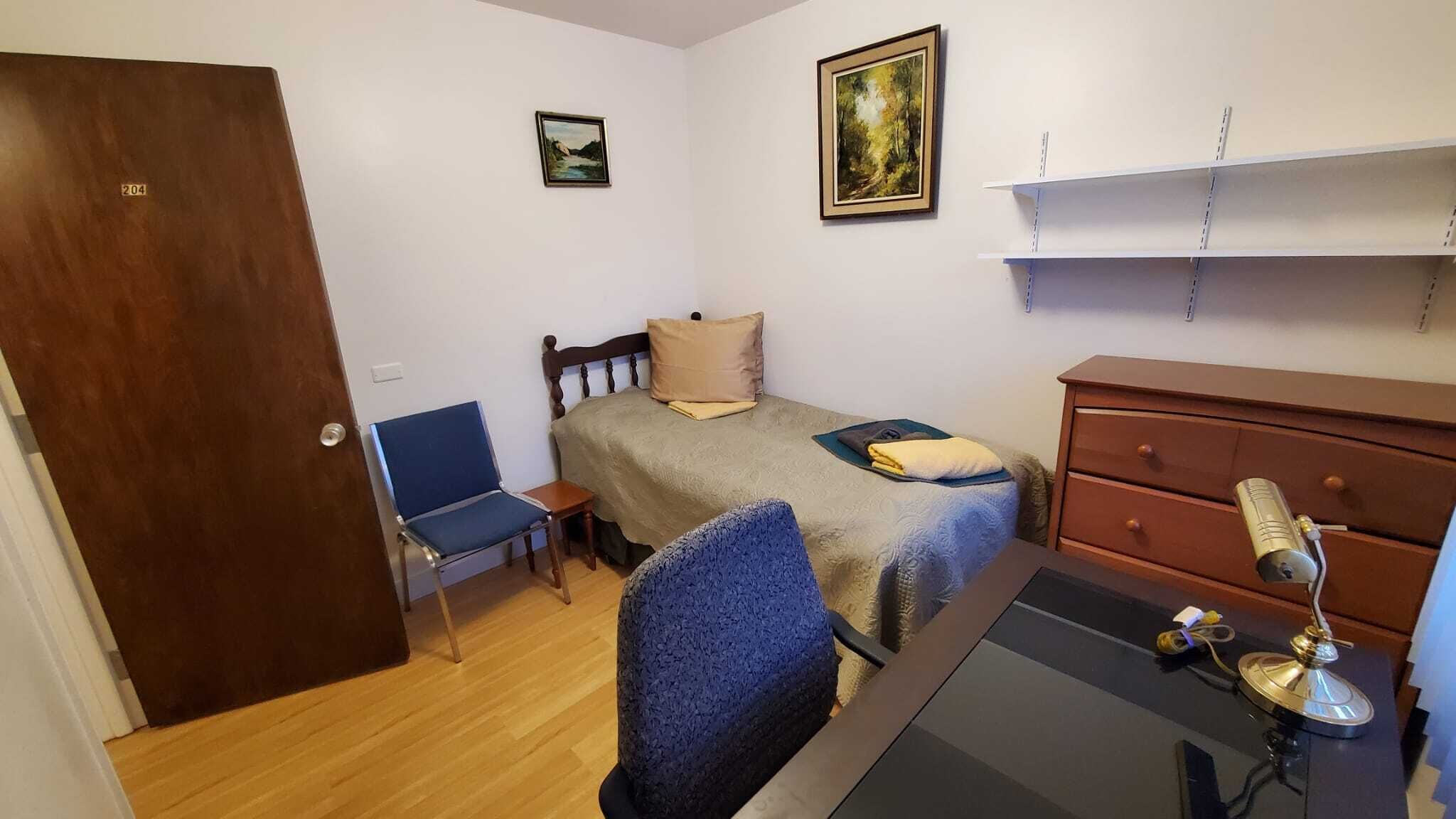 Une petite pièce propre et bien rangée, avec un lit, une chaise, une commode et un bureau doté d’une lampe.