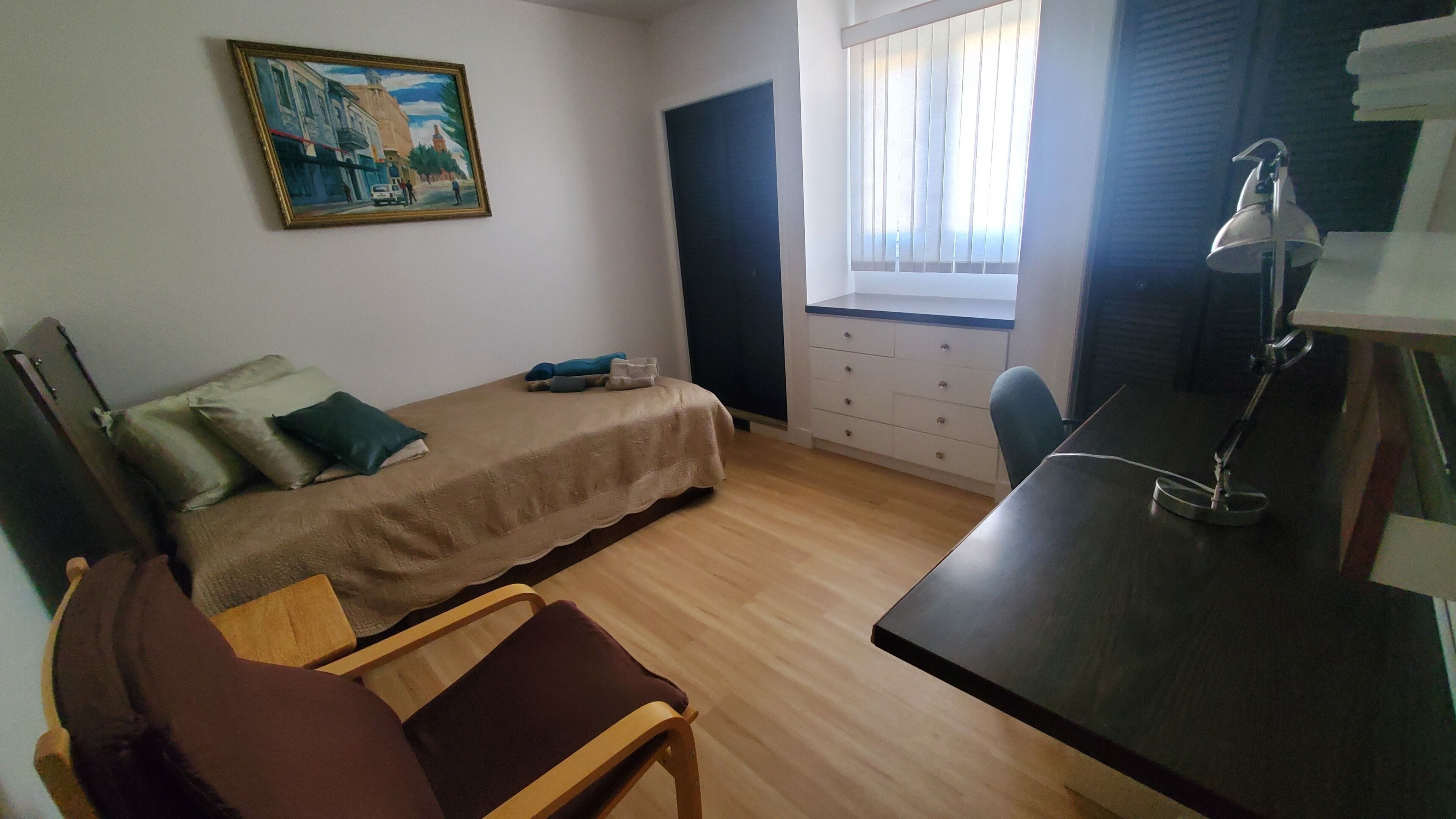 Una habitación pequeña y poco amueblada con una cama, un escritorio, una silla, una ventana con persianas y un cuadro en la pared.