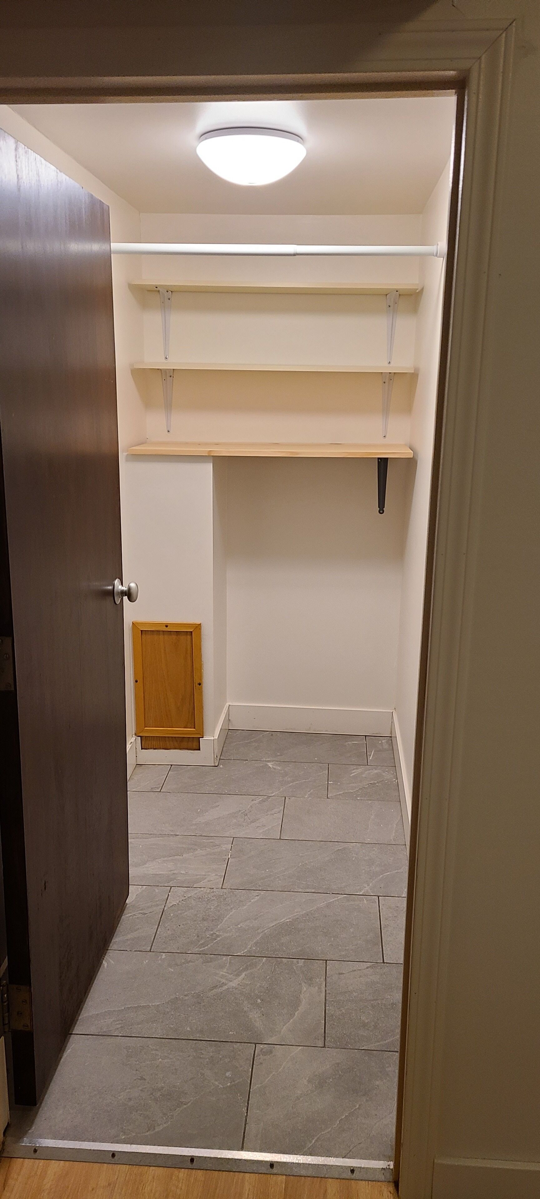 Una habitación con puerta, una lámpara en el techo, una puerta de madera, un armario pequeño de madera y piso de baldosas.
