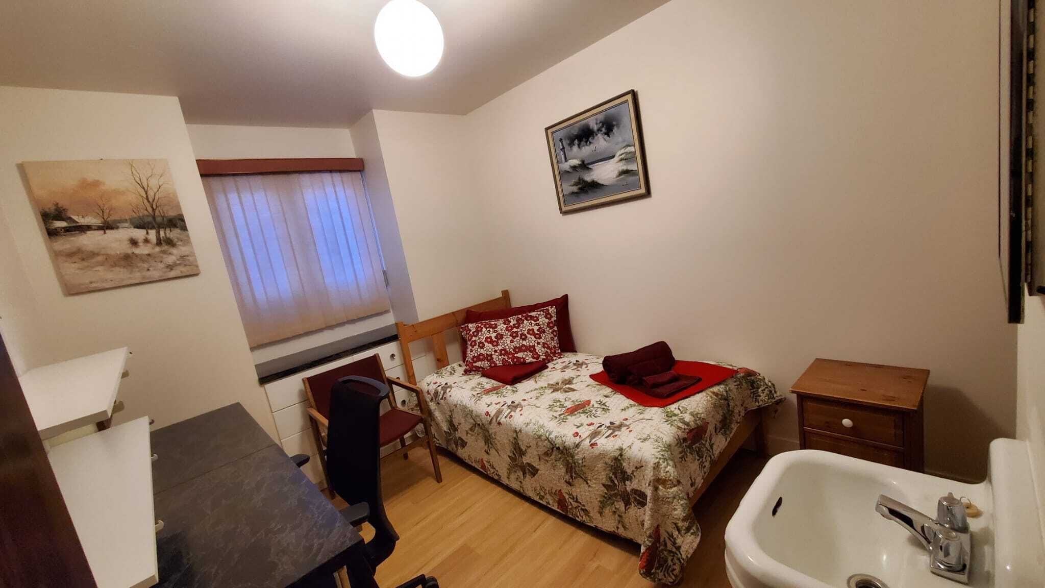 Habitació petita i ben il·luminada amb un llit, escriptori, cadira, finestra amb cortines, un quadre i un rentapans.