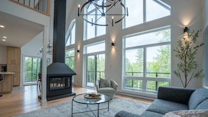A spacious living room with a fireplace, large windows, and a modern chandelier. - New Construction | Spa & Pool Table | Le Foresti (Saint-Alphonse-Rodriguez)