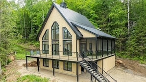 A cabin with a large bay window, a covered deck, and a staircase leading to the main entrance. - New Construction | Spa & Pool Table | Le Foresti (Saint-Alphonse-Rodriguez)