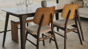 Una mesa de comedor con cuatro sillas y cuatro copas de vino, una planta en maceta y vistas de un edificio a través de grandes ventanales.