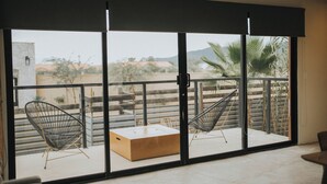 Un balcón con dos sillas de metal y una mesa de madera, con vistas a una zona residencial con palmeras y una montaña distante.