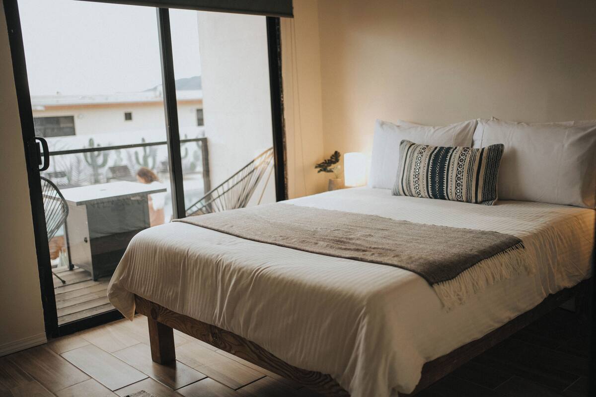 A bedroom with a bed, a nightstand, a lamp, and a view of a balcony.