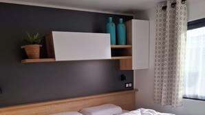 Un dormitorio moderno con cabecera de madera, cama grande con ropa blanca, mesita de noche y estante con objetos decorativos.