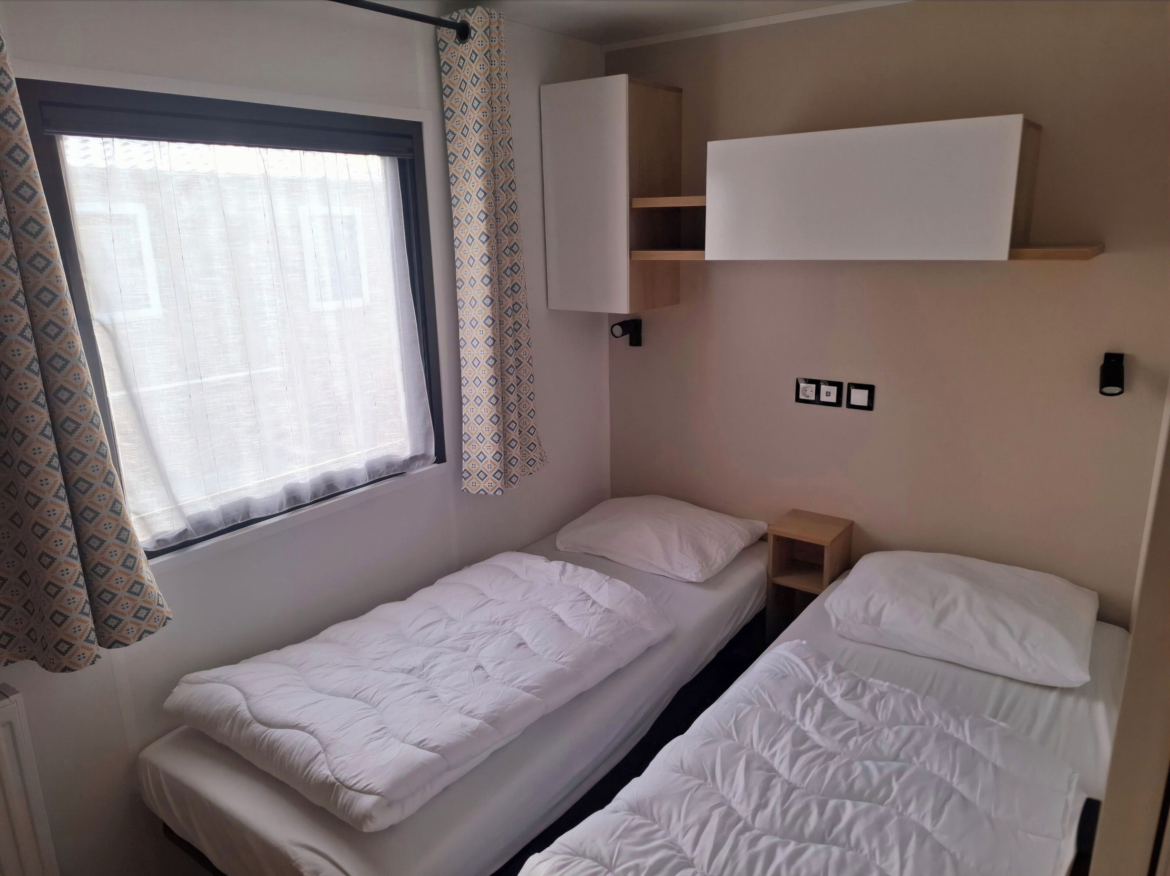A small, clean room with two single beds, a window with curtains, and a wall-mounted shelf.
