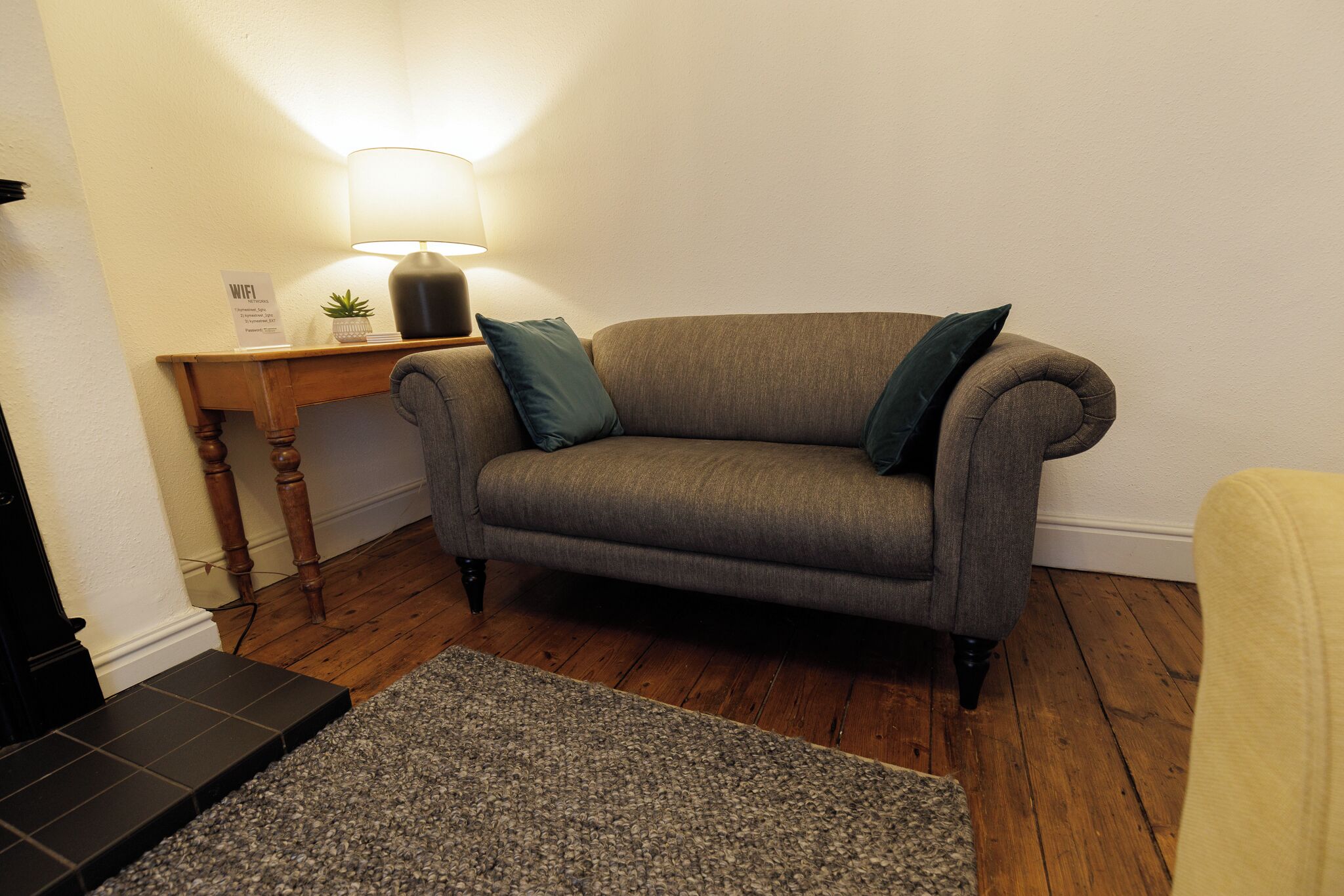 Una sala de estar con un sofá gris, una pequeña mesita de madera, una lámpara y una alfombra gris.