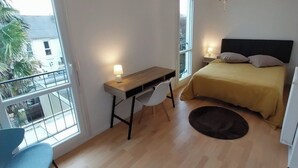 Quarto com cama, mesa de cabeceira, escrivaninha, cadeira e vista para o exterior.