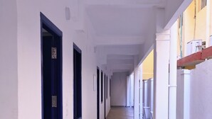 A long, narrow corridor with white walls and blue doors, leading to a building with a tiled floor and a small outdoor area with steps. - Hotel Continental (Puducherry)
