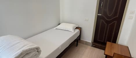 A small, clean room with a single bed, a wooden nightstand, and a door.