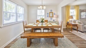 A dining room with a wooden table, chairs, a bench, a chandelier, and a sofa in a bright room with large windows. - Great for Families! Short Drive to Beaches! (Cape May)