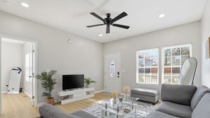 Una sala moderna con un sofá gris, un televisor de pantalla plana sobre un mueble blanco, un ventilador de techo y ventanas grandes con persianas.