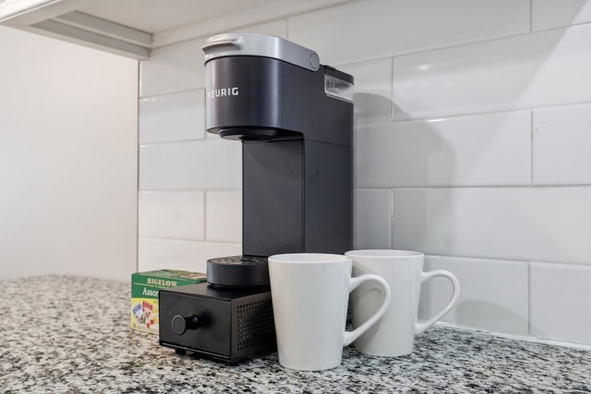 Una cafetera negra con un panel de control y dos tazas blancas sobre una encimera de granito.