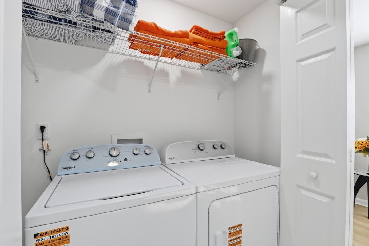 A laundry area with two Whirlpool washers, a metal shelf above holding folded towels and a green bottle, and a white door to the right.