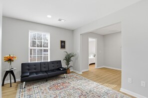 A modern living room with a black sofa, a patterned rug, a potted plant, and a window with blinds. - 4 Bedroom House Perfect for Large Groups Sleeps 10 (Greenville)