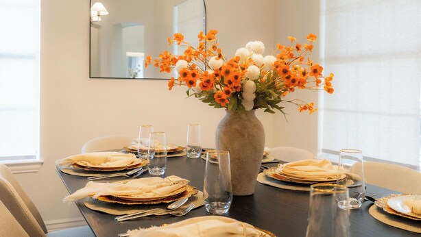 Una mesa de comedor dispuesta con platos, cubiertos y vasos, un florero con flores, y un espejo en la pared.