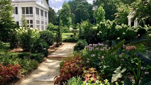A garden pathway leading to a white building with columns, surrounded by lush greenery and flowering plants. - The Home Collection: Modern Myers Park 2BR Condo · Free Parking · Near Uptown & Atrium (Charlotte)