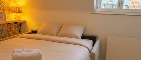 A neatly made bed with rolled towels, a lamp, and a window with a city view.
