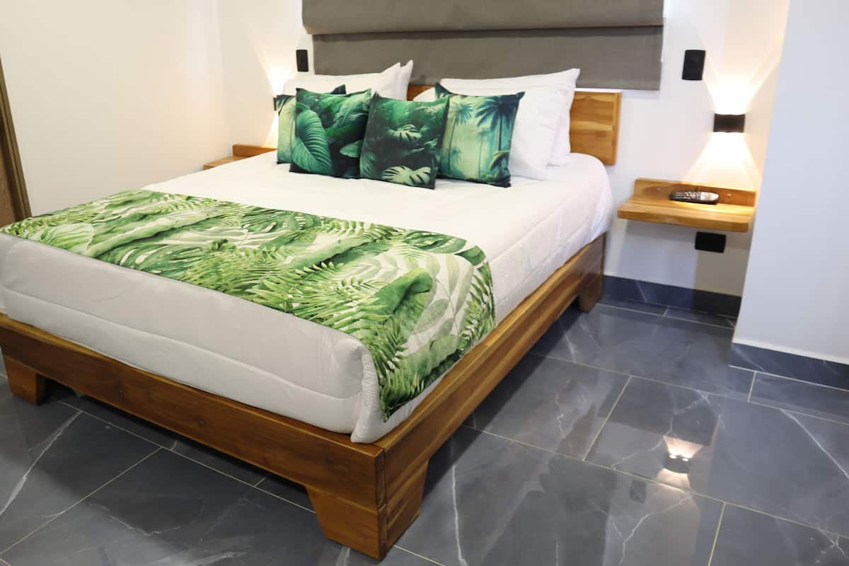 Una cama bien hecha con una colcha de estampado de hojas verdes y almohadas blancas, un cabecero de madera y una mesita de noche con un control remoto.