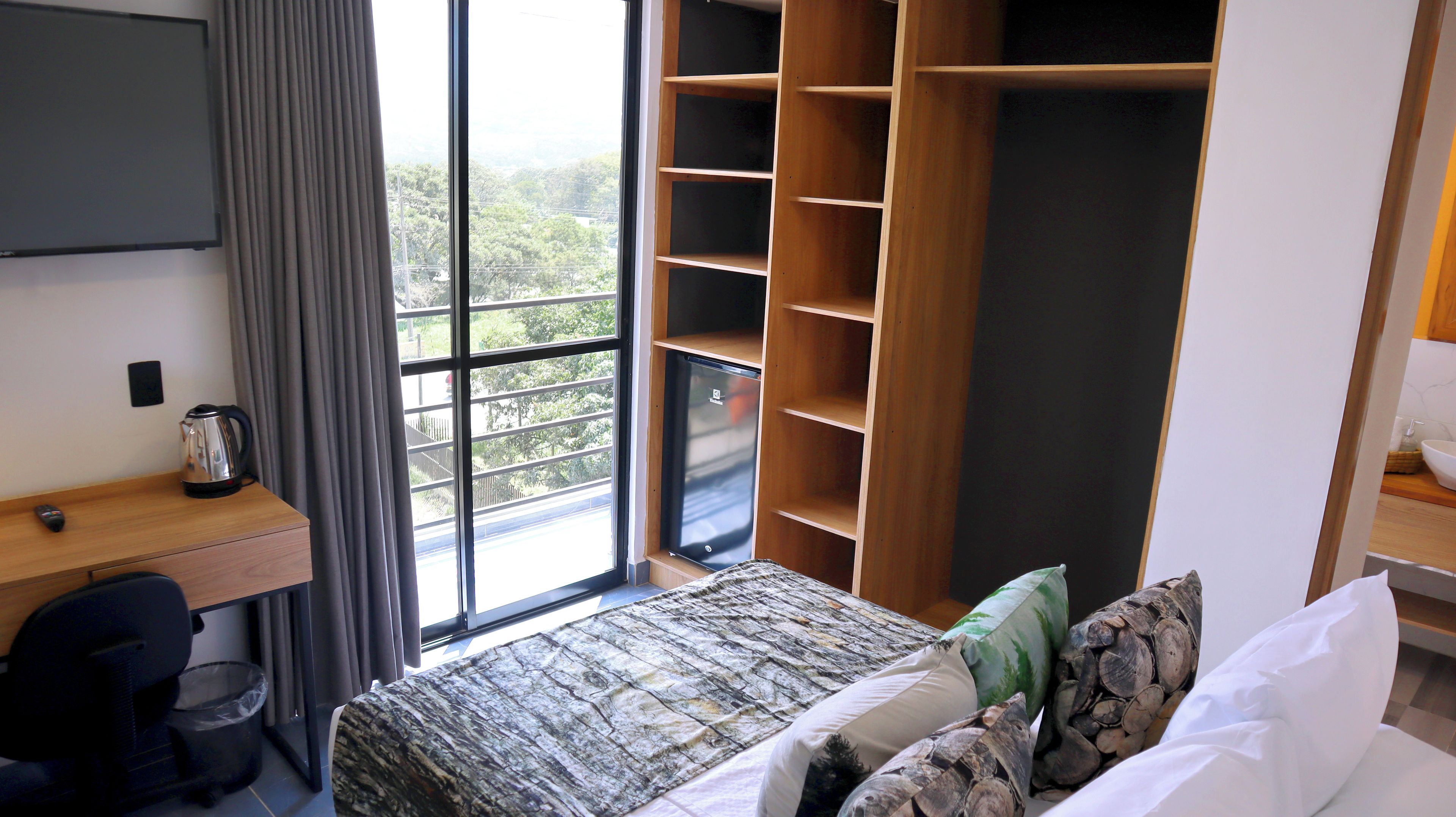 A hotel room with a bed, a desk with a kettle, a wardrobe, and a view of greenery through the window.