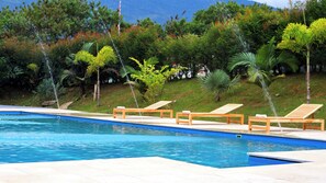 Une piscine avec des chaises longues en bois et une pente herbeuse, avec des arbres en arrière-plan.