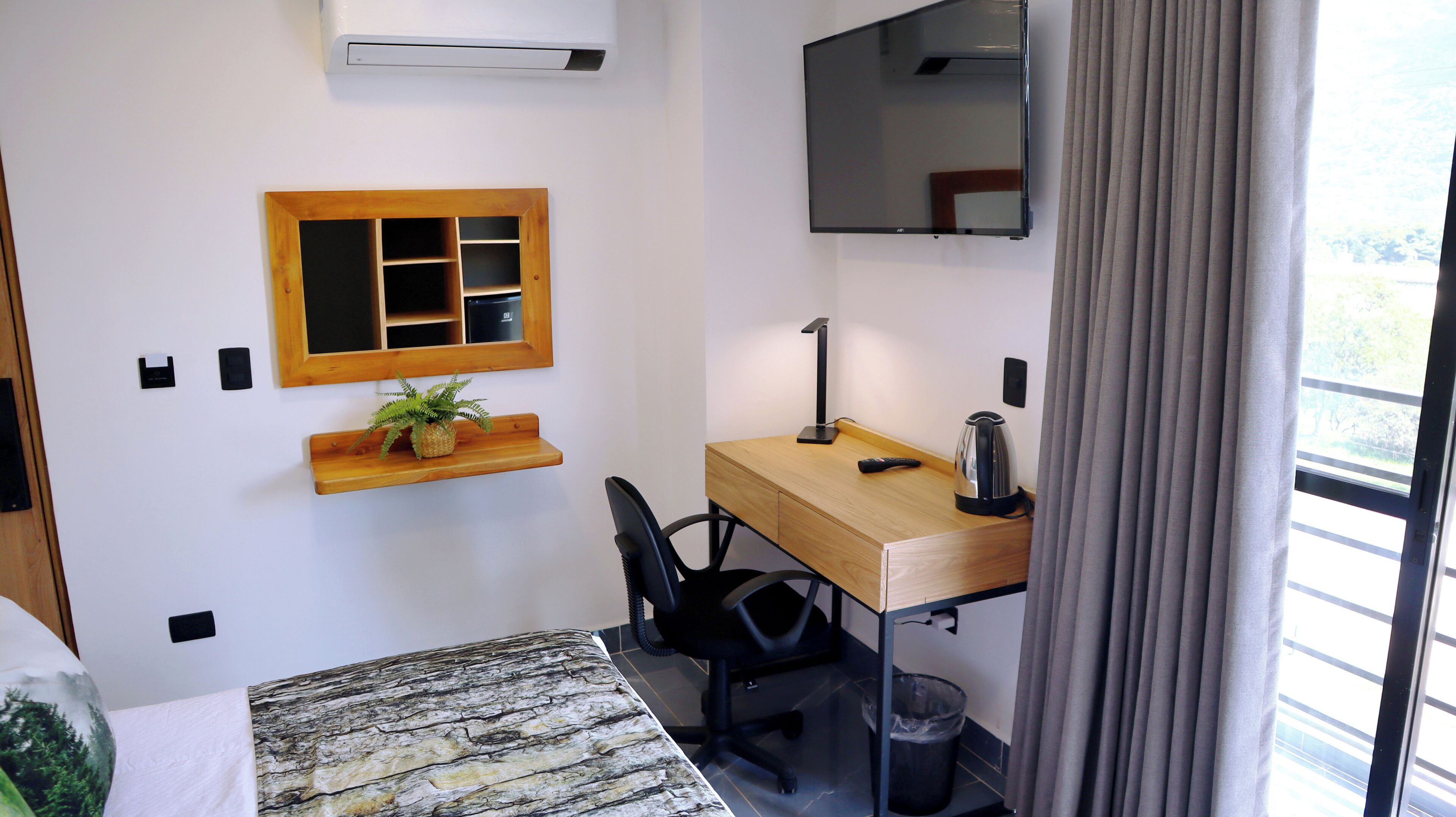 A compact room with a wooden desk, a chair, a small shelf with a plant, a mounted TV, and a window with curtains.