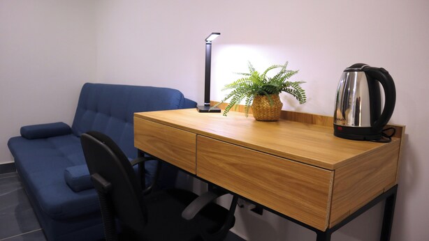 A small office space with a blue chair, a wooden desk with drawers, a black lamp, a potted plant, and a kettle. - GIO GREEN HOTEL GIRARDOTA MEDELLIN (Girardota)