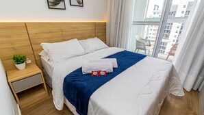 Ein Hotelzimmer mit einem Bett, einem Nachttisch mit Pflanze und Blick auf die Stadt durch große Fenster.