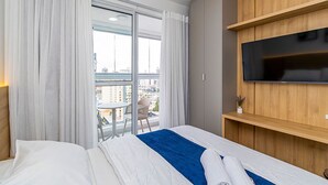 Ein Hotelzimmer mit Bett, einem an einer Holzvertäfelung montierten Fernseher, einem Balkon mit Sitzgelegenheit und Blick auf die Stadt.