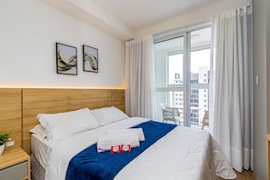 A hotel room with a large bed, a view of the city through a window, and a small balcony with a chair. - Studio com Sacada no Centro - Man1706 (Curitiba)