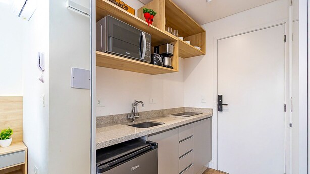 A compact kitchen with a microwave, sink, and dishwasher. There is a wooden shelf above the counter with a potted plant and a coffee maker. - Studio com Sacada no Centro - Man1706 (Curitiba)