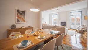 Uma área de refeições com uma mesa de madeira posta para quatro pessoas, um sofá e uma TV ao fundo.