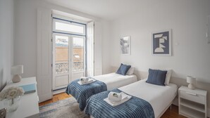 Quarto de hotel com duas camas, uma janela, uma porta e uma pequena mesa com abajur.