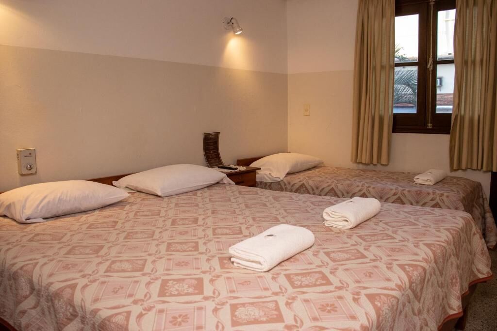 Een hotelkamer met twee bedden, elk voorzien van een beddekleed met een patroon en kussens. Er is een nachtkastje met een lamp en een kleine tafel met een boek. De kamer heeft een raam met gordijnen en uitzicht op het buiten.