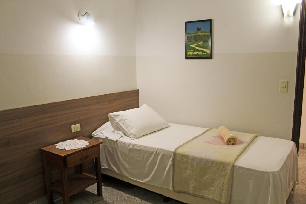 A single bed with a wooden headboard, a small bedside table with a doily, and a poster on the wall.