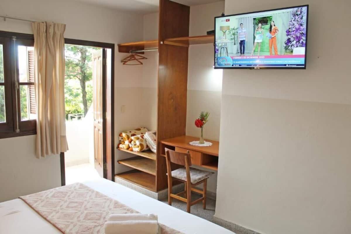 A bedroom with a bed, a desk, a chair, a TV, and a window with curtains.