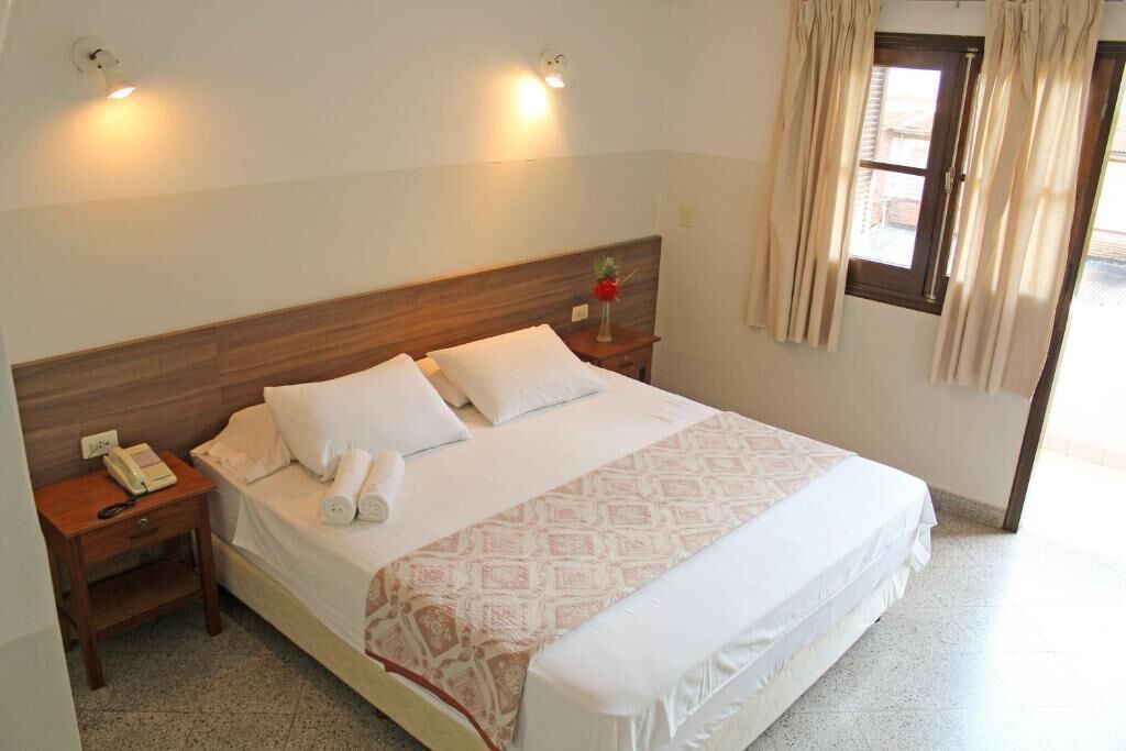 A neatly made bed with white linens and a patterned throw, a bedside table with a lamp and a telephone, and a window with curtains.