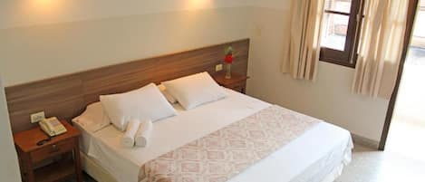 Una cama bien hecha con sábanas blancas y un plaid estampado, una mesita de noche con una lámpara y un teléfono, y una ventana con cortinas.