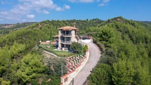 A stone building with a tiled roof, surrounded by lush greenery and a winding road. - Palamidi Apartments (Kassandra)