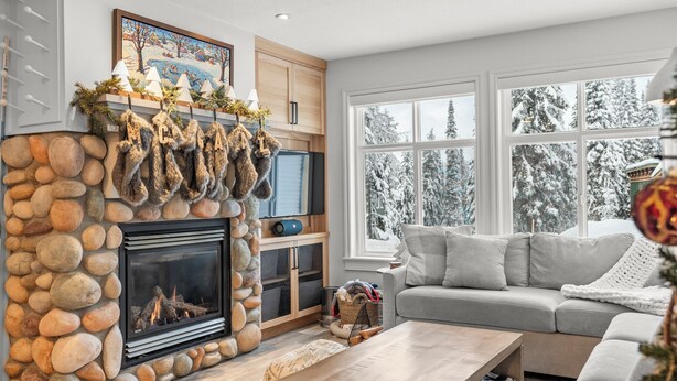 A cozy living room with a stone fireplace, a grey sofa, and a large window with a snowy view. - Modern 2 bedroom ground level suite w/ private hot tub, laundry (Vernon)