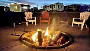 Un foyer avec des bûches, entouré de fauteuils Adirondack, installé sur une terrasse d’où l’on peut admirer les bâtiments en arrière-plan.