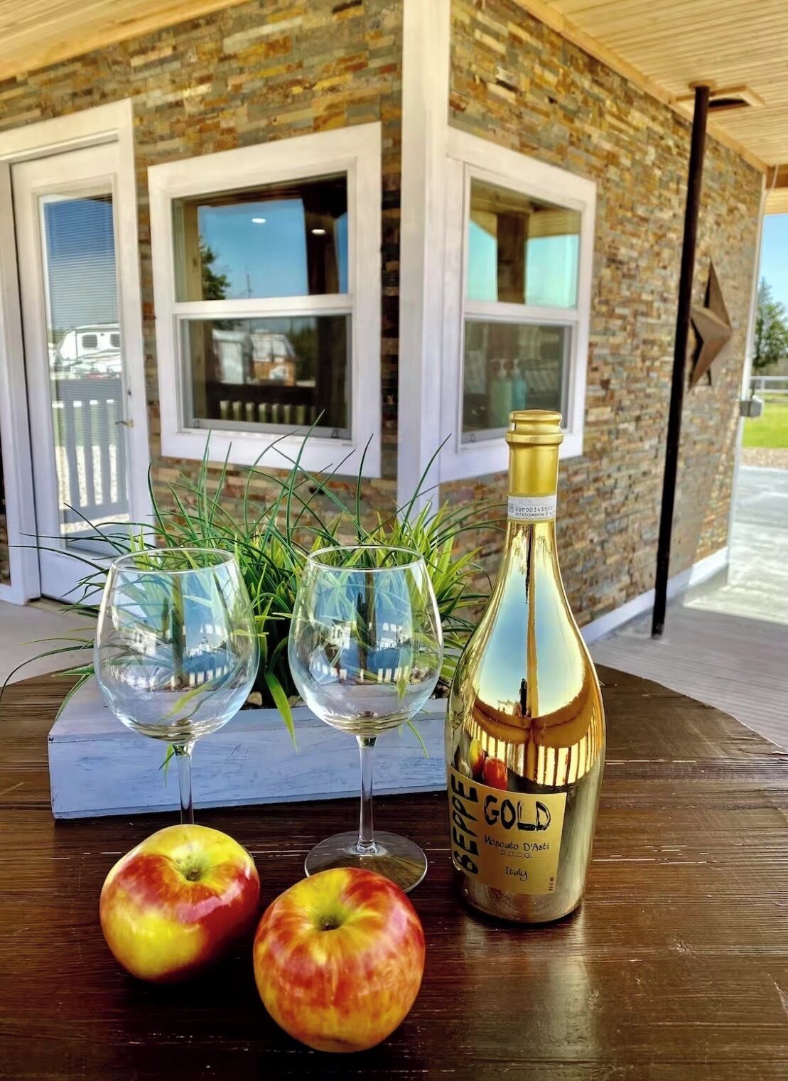 Une table avec deux verres à vin, deux pommes et une bouteille de vin, devant un bâtiment en briques doté de fenêtres.