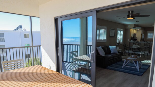 A balcony with a wooden table and chairs, a sliding glass door leading to a living area with a sofa, coffee table, and dining table. - Panoramic Fort Myers Beachfront Condo - Private Sunset Views (Fort Myers Beach)