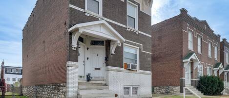 A two-story brick building with a white front door, a small porch, and a stone foundation.